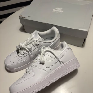 Nike Air Force 1 helvita sneakers - Nike Air Force 1 sneakers i helvit färg med klassisk låg siluett. Skorna har snörning, perforerad tå och rund tå. Tillverkade i skinn med platt sula och ikonisk Nike-logga på sidan och hälen. Perfekta för dig som gillar clean och tidlös streetstil.