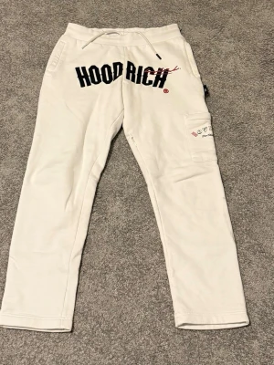 Vita mjukisbyxor från Hoodrich XS - Säljer ett par vita mjukisbyxor från Hoodrich i storlek XS. Byxorna har snörning i midjan, tryck med HOODRICH på framsidan och en ficka med grafiskt tryck på benet. Sköna och stilrena med loose passform, perfekta för chill eller streetwear.