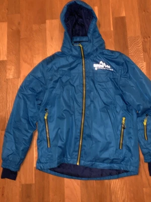 Blå skidjacka från Crivit med huva - Blå skidjacka från Crivit med huva och gula dragkedjor. Jackan har två sidofickor med dragkedja och reflekterande tryck fram och bak. Tillverkad i polyester och har ribbade muddar vid ärmslut. Perfekt för blåsiga dagar och aktivitet som skid sam snowboardåkning 