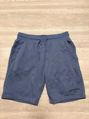 Blå shorts från Björn Borg - Säljer ett par mörkblå shorts från Björn Borg med snörning i midjan och fickor på sidorna samt en bakficka. Mjukt bomullsmaterial som är skönt att ha på sig. Perfekta för chill dagar eller när du vill vara bekväm.