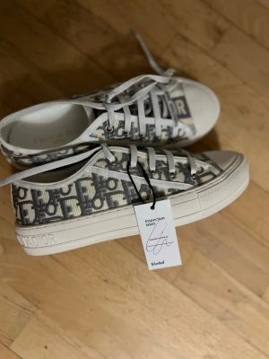 Christian Dior sneakers mönstrade - Säljer ett par snygga Christian Dior sneakers i vit och grå färg med ikoniskt Dior-mönster. Skorna har snörning, rund tå och platt sula med en svart stjärna undertill. Tillverkade i Italien och har textilöverdel med tydlig logga på plösen.