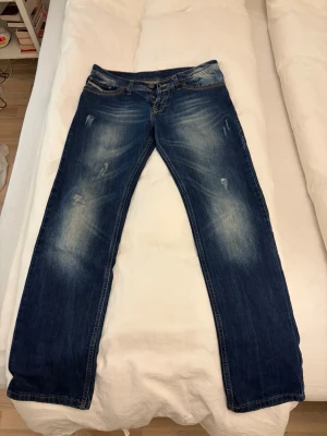Blå Diesel jeans med slitningar - Säljer ett par blå jeans från Diesel med snygga slitningar och tvättade detaljer. Klassisk femficksmodell med bruna sömmar och läderpatch bak. Jeansen har raka ben och normal passform, tillverkade i slitstarkt denim. Perfekt för dig som gillar en cool och avslappnad stil. Pris inte hugget i sten. Tar även byten.