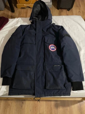 Mörkblå Canada Goose Expedition jacka - Säljer en mörkblå Canada Goose Expedition jacka med huva och flera stora fickor. Jackan har det klassiska Arctic Program-märket på bröstet och är tillverkad i slitstark polyester. Perfekt för kalla vinterdagar och har en loose passform.