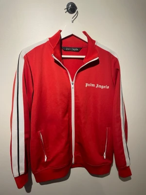Röd Palm Angels track jacket - Palm Angels track jacket i rött med vita och svarta ränder längs ärmarna. Jackan har ståkrage, hel dragkedja framtill och två fickor med dragkedja. Loggan är tryckt på bröstet. Perfekt för en sportig och trendig look.