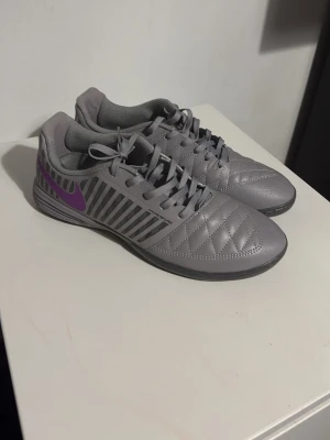 Grå Nike fotbollsskor med lila swoosh - Säljer ett par grå Nike fotbollsskor med snygga ränder på sidorna och en lila swoosh. Skorna har snörning och quiltad ovandel i syntetmaterial, samt platt sula som passar perfekt för inomhusspel. Kommer i originalkartong.