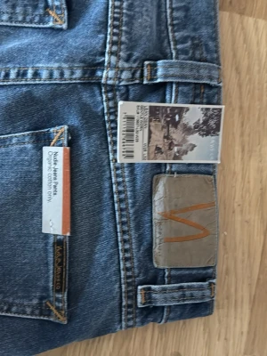 Nudie Jeans blå straight jeans lean Dean  - Säljer ett par klassiska blå jeans från Nudie Jeans, tillverkade i ekologisk bomull. Modellen har raka ben och snygga slitningar för en cool look. Jeansens patch baktill har Nudie-loggan i orange. Perfekt för dig som gillar tidlös stil.