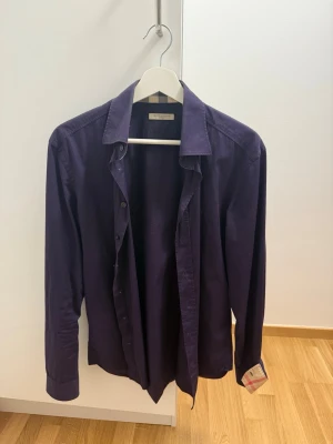 Mörklila skjorta från Burberry - Snygg mörklila skjorta från Burberry i bomull med klassisk krage och knappar framtill. Långärmad modell med en diskret Burberry-rutig detalj på insidan av manschetten. Perfekt för dig som gillar stilrena plagg med en twist.