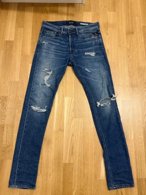 Replay Broken Edge blå jeans, W31 L34 - Säljer ett par Replay Broken Edge jeans i klassisk blå tvätt med slitna detaljer och hål på benen. Modellen har smal passform, fem fickor och dragkedja. Perfekt för dig som gillar en edgy streetstil. Materialet är jeans och de har en snygg mörkblå ton.
