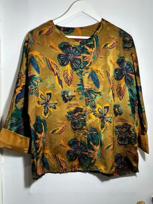 Silkesjacka - Elegant jacka i 100 % siden med ett rikt, måleriskt blommönster i varma gröna och gyllene toner. Tyget är mjukt, lätt och följsamt med en naturlig lyster som ger plagget ett exklusivt uttryck.  Avslappnad passform som faller fint på kroppen. De dekorativa knäppningarna framtill ger en subtil orientalisk känsla och gör jackan både personlig och tidlös.  🧵 Material 	•	100 % siden  📐 Passform 	•	Bekväm, något rymlig modell 	•	Lätt att bära både som tunn jacka eller som överdel  Perfekt att kombiner