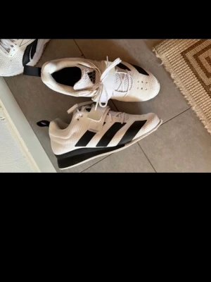 Vita och svarta Adidas träningsskor - Säljer ett par vita Adidas träningsskor med svarta ränder på sidorna. Skorna har snörning och en extra rem över vristen för bättre passform. Ovandelen är i ventilerande syntetmaterial och sulan är platt och stabil. Perfekta för gym eller sport.