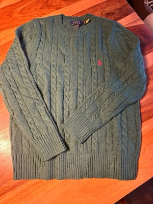 Grön Polo Ralph Lauren tröja (storlek M) - Snygg grön kabelstickad tröja från Polo Ralph Lauren med klassisk rund hals och långärmad design. Tröjan har ett broderat rosa Polo-logo på bröstet och ribbade muddar vid ärmslut och nederkant. Perfekt för dig som gillar stilren och tidlös look.