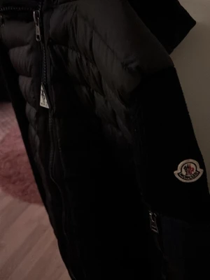 Svart cardigan från Moncler - Svart cardigan från Moncler med huva och ribbade muddar. Jackan har dragkedja framtill, två sidofickor med en knapp och ett Moncler-märke på ärmen. Materialet är polyester och dun, perfekt för kyliga dagar. Snygg och stilren design som funkar till många outfits. Jag har inte använt den mycket så den är i bra skick. Skicka om ni är intresserade!