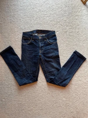 Mörkblå slim fit jeans från Nudie - Snygga mörkblå jeans från Nudie med klassisk femficksdesign och smal passform. Jeansen har en läderpatch med Nudie-logga bak och är tillverkade i slitstarkt bomullsmaterial. Perfekta för dig som gillar en tight och modern look.