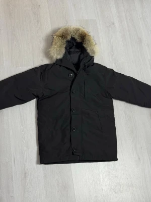  Canada Goose Chateau  - Svart parkajacka från Canada Goose med fluffig päls runt huvan och snygga detaljer som knappar och dragkedja. Jackan har lång ärm, och en varm vinterjacka. Säljs pågrund av fel storlek men annars är den jätte fin, lappar finns kvar på jackan. 