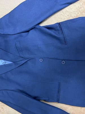 Blå kavaj med två knappar allt inklusive  - Snygg blå kavaj med klassisk passform och två knappar framtill. Jackan har smala slag, raka fickor och fyra dekorativa knappar vid ärmslutet. Perfekt för dig som vill ha en stilren look med lite färg.