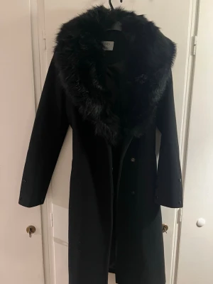 Svart kappa med fuskpäls och bälte - Säljer en svart lång kappa från Hollies med bred krage och muddar i fuskpäls. Kappan har bälte i midjan och knappar framtill. I bra skick , inga defekter . Storlek 34. Pälsen är avtagbara och kappan kan användas utan också .