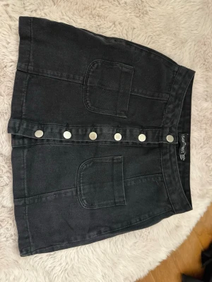 Svart kortkjol från I Saw It First - Säljer en svart kortkjol från I Saw It First med två stora fickor framtill och snygga silvriga knappar längs hela framsidan. Kjolen är i ett stadigt bomullsmaterial och har en rak passform som ger en cool och enkel look. Kjolen är lite urtvättad i färgen