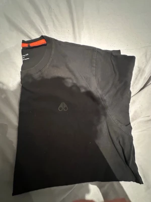 Svart t-shirt Moose Knuckles L - Svart t-shirt från Moose Knuckles i storlek L, tillverkad av 100% ekologisk bomull. Diskret logga på bröstet och tryckt varumärke vid nederkanten. Snygg orange detalj på insidan av kragen. Perfekt för dig som gillar stilrena och bekväma plagg.