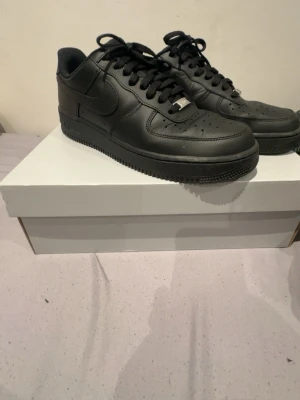Nike Air Force 1 helsvarta sneakers - Säljer ett par klassiska Nike Air Force 1 i helsvart skinn. Skorna har låg profil, rund tå, snörning och den ikoniska tjocka sulan. Perfekta för dig som vill ha en clean och tidlös look. Passar till allt och är riktigt bekväma.
