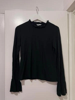 Svart tröja med volangkrage - Säljer en svart tröja från cubus med långärmade ärmar och volangdetaljer vid kragen och ärmsluten. Tröja har en klassisk, lite loose passform och är tillverkad i mjuk polyester. Perfekt för dig som gillar stilrena och feminina detaljer.