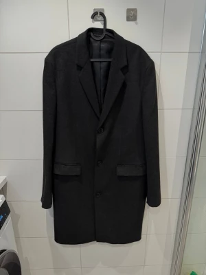 Klassisk rock från Zara - strl XL - Mörkgrå rock med klassisk krage och tre knappar framtil. Jackan har två diskreta fickor och är helfodrad på insidan. Passformen är rak och ärmarna är långa med knappar vid ärmslutet. Perfekt för dig som gillar stilrena och tidlösa plagg.