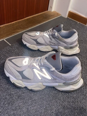 New Balance 9060 grå sneakers - Säljer ett par New Balance 9060 sneakers i grått med vita och mörkgrå detaljer. Skorna har en chunky sula, meshpaneler och mockadetaljer för en modern look. Reflekterande N-logga på sidan och snörning framtill. Perfekta för dig som gillar streetwear och bekväma skor. Ifall du vill att jag ska rengöra skorna säg bara till. Tack mvh
