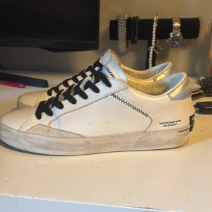 Vita sneakers från Crime London - Snygga vita sneakers från Crime London med svarta snören, silvriga detaljer på hälen och en orange accent på sidan. Skorna har textdetaljer och coola kontrastsömmar. Tillverkade i skinn med platt sula och rund tå. Perfekta för dig som gillar streetstyle med edge.