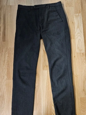 Svarta slim kostymbyxor Jack&Jones 28/32 - Snygga svarta/mörkgråa kostymbyxor från Jack&Jones i slim fit-modell. Byxorna har klassiska fickor fram och bak, bälteshällor och knappstängning. Perfekt för dig som gillar stilrena och smala byxor. Passar dig som vill ha en modern look eller till kostym.