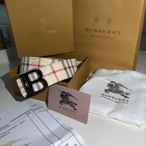 Burberry bälte med B-spänne - Säljer ett Burberry bälte med ikoniskt rutigt mönster i beige, rött och svart samt ett stort B-spänne i metall. Bältet är tillverkat i skinn och har klassisk Burberry-stil. Perfekt accessoar för att lyfta din outfit. Kom med bud hundra procent accepterar!!!