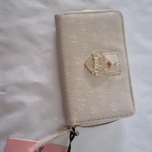Vit plånbok från Juicy Couture - Superfin vit plånbok från Juicy Couture med präglat JC-mönster och guldfärgad hjärt-detalj framtill. Plånboken har dragkedja runtom, flera kortfack, ID-fönster och texten 'My Heart Belongs to Juicy' i guld. Insidan har coolt Juicy Couture-tryck.