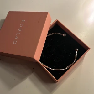 Silvrigt armband från Edblad - Snyggt armband från Edblad i silverfärgad metall med tvinnad design och två runda kulor i ändarna. Kommer i original ask, perfekt för dig som gillar stilrena och moderna accessoarer.