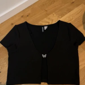 Svart croppad bolerotopp från H&M Divided - Svart croppad bolerotopp från H&M Divided med korta ärmar. Toppen är ribbad och har en söt silverfärgad fjärilsdetalj som knäppning framtill. Perfekt att styla över linne eller klänning för en trendig look.