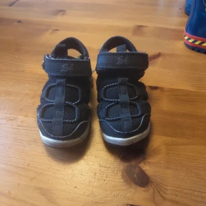 Svarta sandaler från Leaf, strl 23 - Säljer ett par svarta sandaler från Leaf i storlek 23. De har sportig design med justerbar kardborreknäppning och robust sula. Ovandelen är i textil och syntet, perfekt för aktiva dagar under sommaren. Sulan blinkar