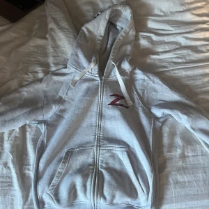 Grå Zorro hoodie med tryck - Snygg grå hoodie med dragkedja och huva ifrån russemerch . På bröstet finns ett rött Z-tryck och på ryggen ett stort Zorro-motiv i rött och svart samt texten 'BECKER'. Mjuk insida och kängurufickor framtill. Perfekt för dig som gillar streetstyle.
