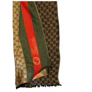 Gucci halsduk med GG-mönster - Snygg halsduk från Gucci med klassiskt GG-mönster i beige och mörkbrunt. Har gröna och röda ränder med Gucci-logga längs kanten. Tunn och mjuk, perfekt att slänga över axlarna för en lyxig touch. Fransad kant för extra stilpoäng.