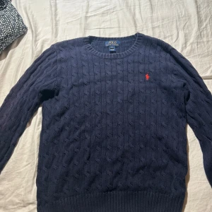 Mörkblå kabelstickad tröja Polo Ralph Lauren - Kabelstickad mörkblå tröja från Polo Ralph Lauren med rund halsringning och klassisk röd logga broderad på bröstet. Tröjan har långa ärmar och ribbade muddar. Perfekt för dig som gillar en stilren och tidlös look.  14-16 år