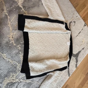 Beige kofta med svarta detaljer och guldknappar - Säljer en beige kofta med svarta kanter och snygga guldknappar framtill. Koftan har lång ärm och är tillverkad i ett strukturerat, stickat material som känns både trendigt och klassiskt. Perfekt för dig som vill ha en stilren och chic look.