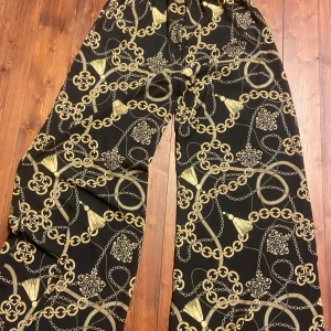 Svarta vida byxor med kedjemönster - Snygga svarta byxor med guldigt kedjemönster och vida ben. Byxorna har en riktigt trendig look med sitt iögonfallande print och passar perfekt för dig som vill sticka ut. Materialet känns mjukt och följsamt mot huden.