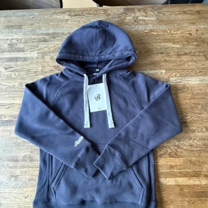Mörkblå hoodie från Russemerch - Mörkblå hoodie från Russemerch med vit snörning i huvan och broderad logga på ärmen. Tröjan har känguruficka framtill och är tillverkad i mjuk bomullsblandning. Perfekt för chill dagar och enkel att matcha med jeans eller mjukisbyxor. OBS⚠️aldrig använt fick hem den idag och testade den, den va för liten för mig helt ny. Etiketten är kvar och helt ny