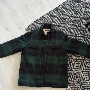 Grönrutig overshirt med teddyfoder - Snygg overshirt i mörkgrönt och svart rutigt mönster med mjukt, ljust teddyfoder på insidan. Jackan har två bröstfickor med lock, knäppning framtill och klassisk krage. Perfekt för lager-på-lager och chill höst- eller vinterstil. Använd 1 gång.