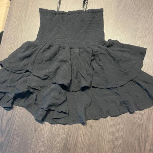 Svart volangkjol med smock - Supersöt svart kortkjol med bred smock i midjan och dubbla volanger. Kjolen har en luftig känsla och passar perfekt till en trendig look. Materialet känns lätt och flowy, vilket gör den extra bekväm.