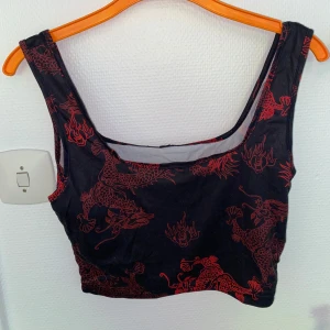 Svart crop top med röda drakar - Cool svart croppad topp med röda drakmönster. Toppen är ärmlös och har breda axelband samt rundad halsringning. Perfekt för dig som vill sticka ut med ett asiatiskt inspirerat print. Materialet känns mjukt och stretchigt.