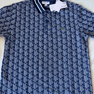 Lacoste Piké Polo  - Snygg blå pikétröja från Lacoste med vitt geometriskt mönster över hela tröjan. Klassisk krage med vita ränder och knäppning framtill. Ikoniska gröna krokodilloggan på bröstet. Perfekt för dig som gillar stilrena detaljer och sportig vibe.