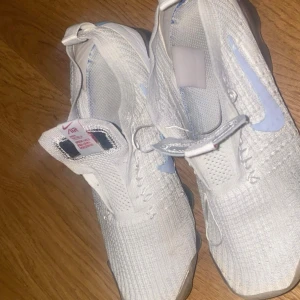 Nike Air Vapormax Flyknit vita sneakers - Nike Air Vapormax Flyknit sneakers i vitt med ljusblå swoosh. Skorna har en stickad ovandel i flyknit-material och genomskinlig sula. De har en modern design med kardborreband och är superlätta och bekväma för vardagsbruk. 