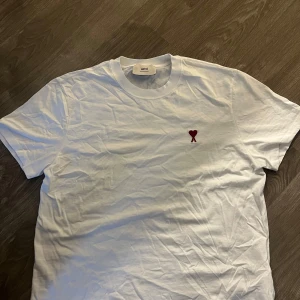Vit t-shirt från Ami med rött hjärta - Vit t-shirt från Ami Alexandre Mattiussi med klassisk passform och rund hals. På bröstet finns ett broderat rött hjärta med bokstaven A. Tillverkad i mjuk bomull, perfekt för en clean och stilren look.