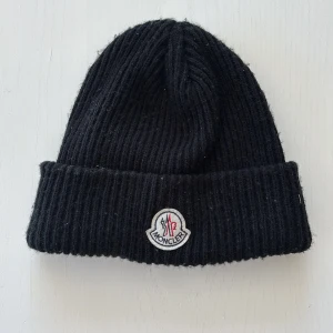 Svart Moncler mössa i ull - Svart ribbstickad mössa från Moncler med uppvikt kant och klassisk Moncler-logga framtill. Tillverkad i mjuk ull som håller dig varm under kyliga dagar. Enkel och stilren design som passar perfekt till streetwear och vinteroutfits.