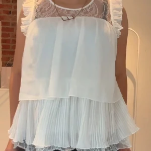 Vit volangblus med spetsdetaljer - Supersöt vit blus med volanger på axlarna, plisserad nederdel och transparent spets upptill. Blusen är ärmlös och har en luftig, flowy passform som känns både romantisk och trendig. Perfekt för dig som gillar feminina detaljer och lager på lager.