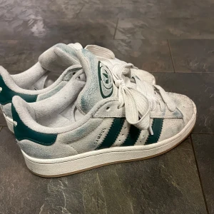 Adidas Campus sneakers vit/grön mocka - Adidas Campus sneakers i vit mocka med gröna detaljer och klassiska tre ränder på sidan. Skorna har rund tå, platt sula och breda vita skosnören. Adidas-logga på plösen och hälen. Perfekta för dig som gillar retrostil och streetwear.