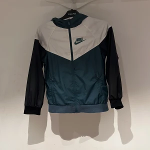 Nike vindjacka i blått och vitt - Nike vindjacka med huva i blått, vitt och svarta ärmar. Jackan har dragkedja framtill, två fickor med dragkedja och meshfoder. Snygg sportig look med klassisk Nike-logga på bröstet. Perfekt för dig som gillar streetwear och sportig stil.