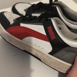 Puma sneakers i svart, vitt och rött - Snygga Puma sneakers i storlek 39 med klassisk design. Skorna har svart, vitt och grått skinn med den ikoniska röda Puma-loggan på sidan. De har svarta snören och en vit sula med Puma-tryck. Perfekta för dig som gillar sportig och stilren look.